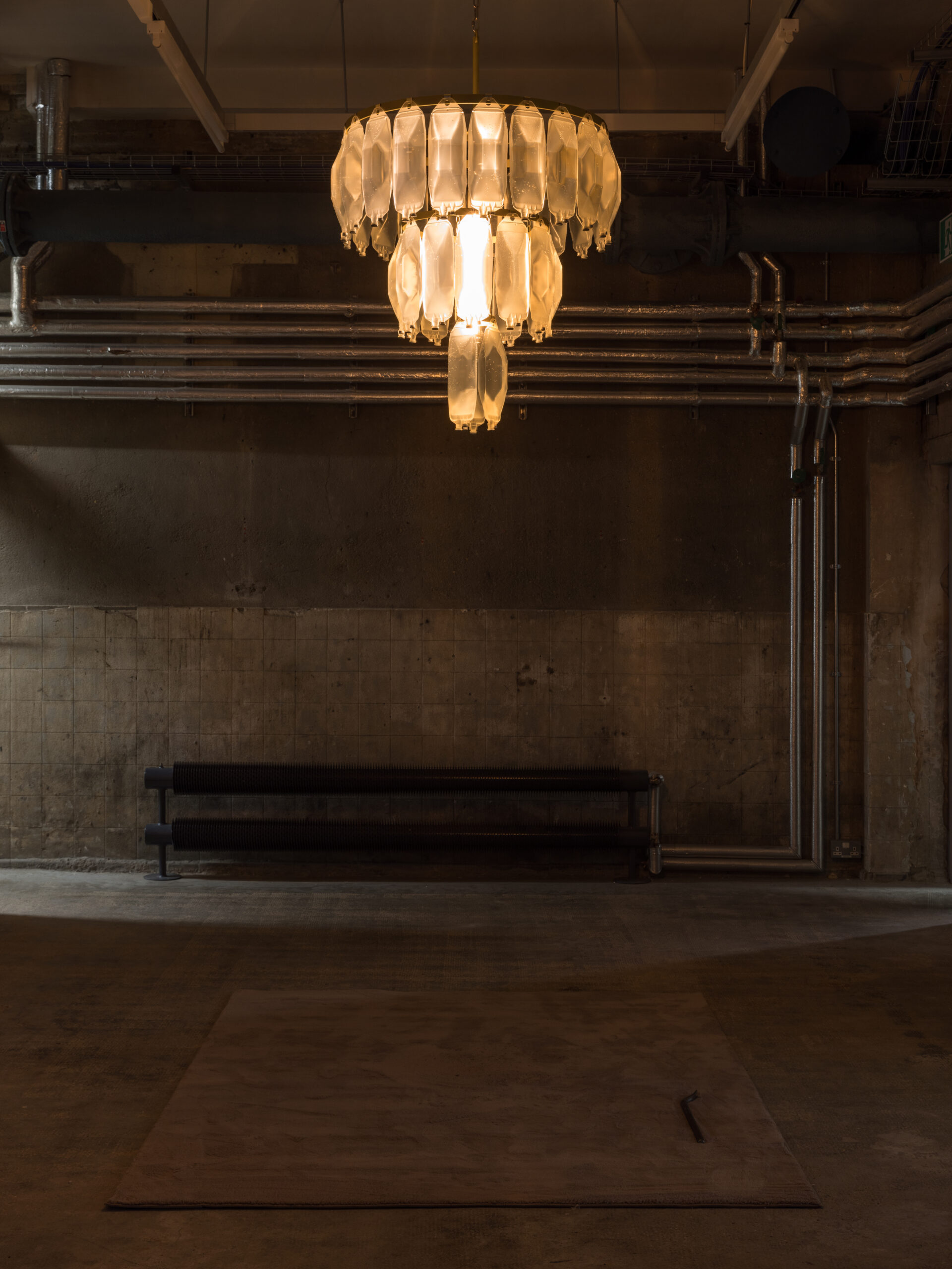 Avril Corroon, Sublet Glory, 2026, dehumidifier water (collected from London tenancies), IV bags, crowbar, steel, brass, electric components, carpet, installation view, ‘Flare-Up’ (21 May –16 August 2026) at Goldsmiths Centre for Contemporary Art; photograph by Rob Harris, courtesy of the artist and Goldsmiths CCA.